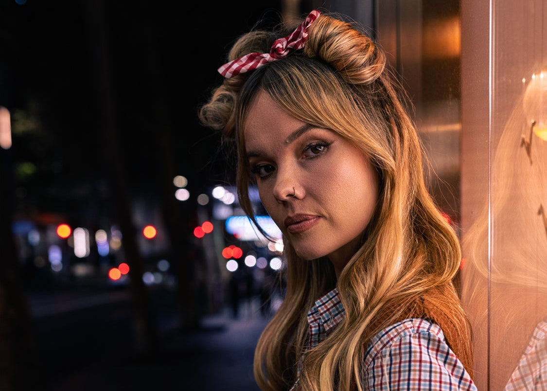 Woman in a plaid shirt and brown vest standing against a dark background